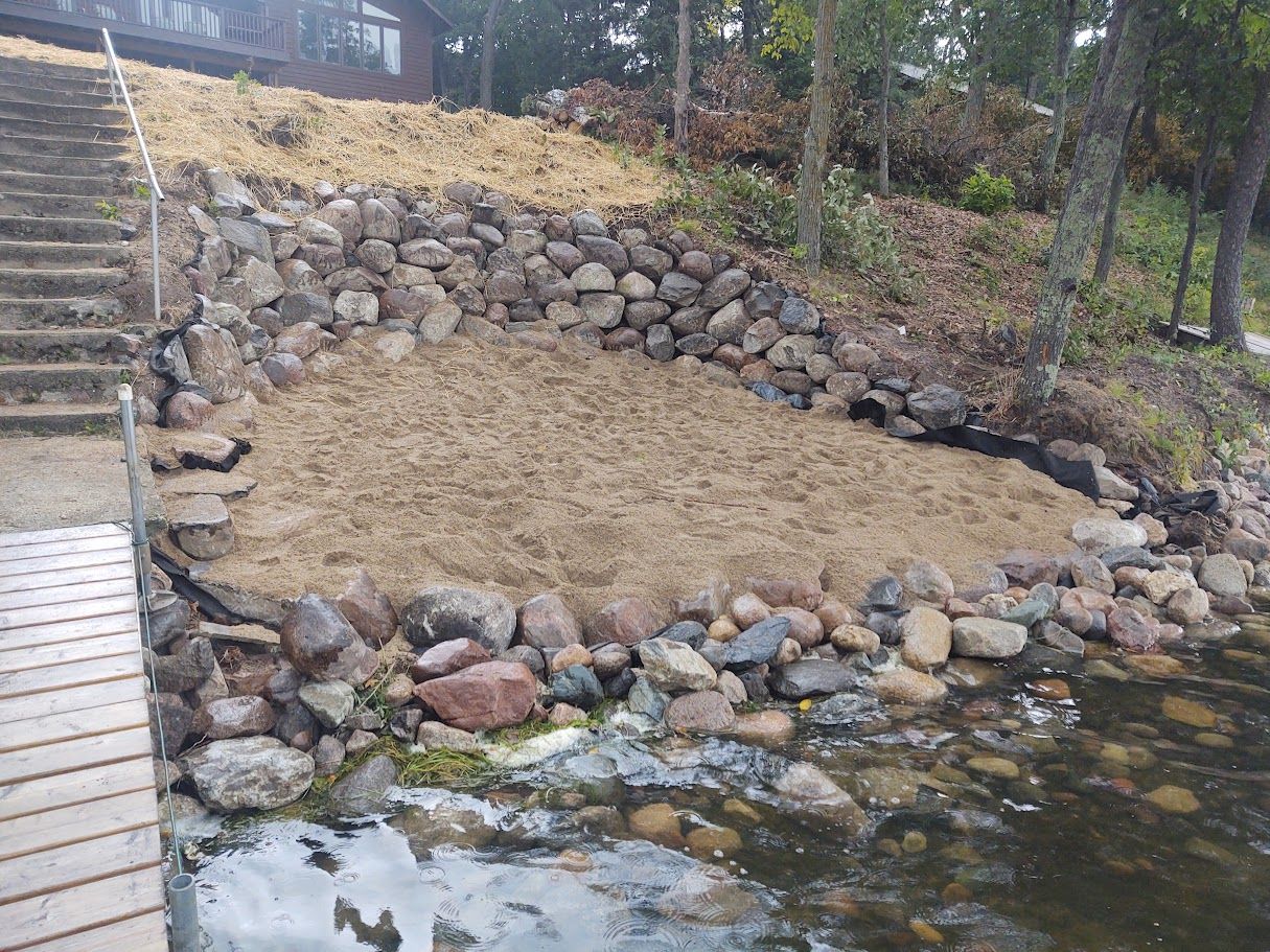 Rock retaining wall around a bare sandy slope beside water, with stone steps on the left.