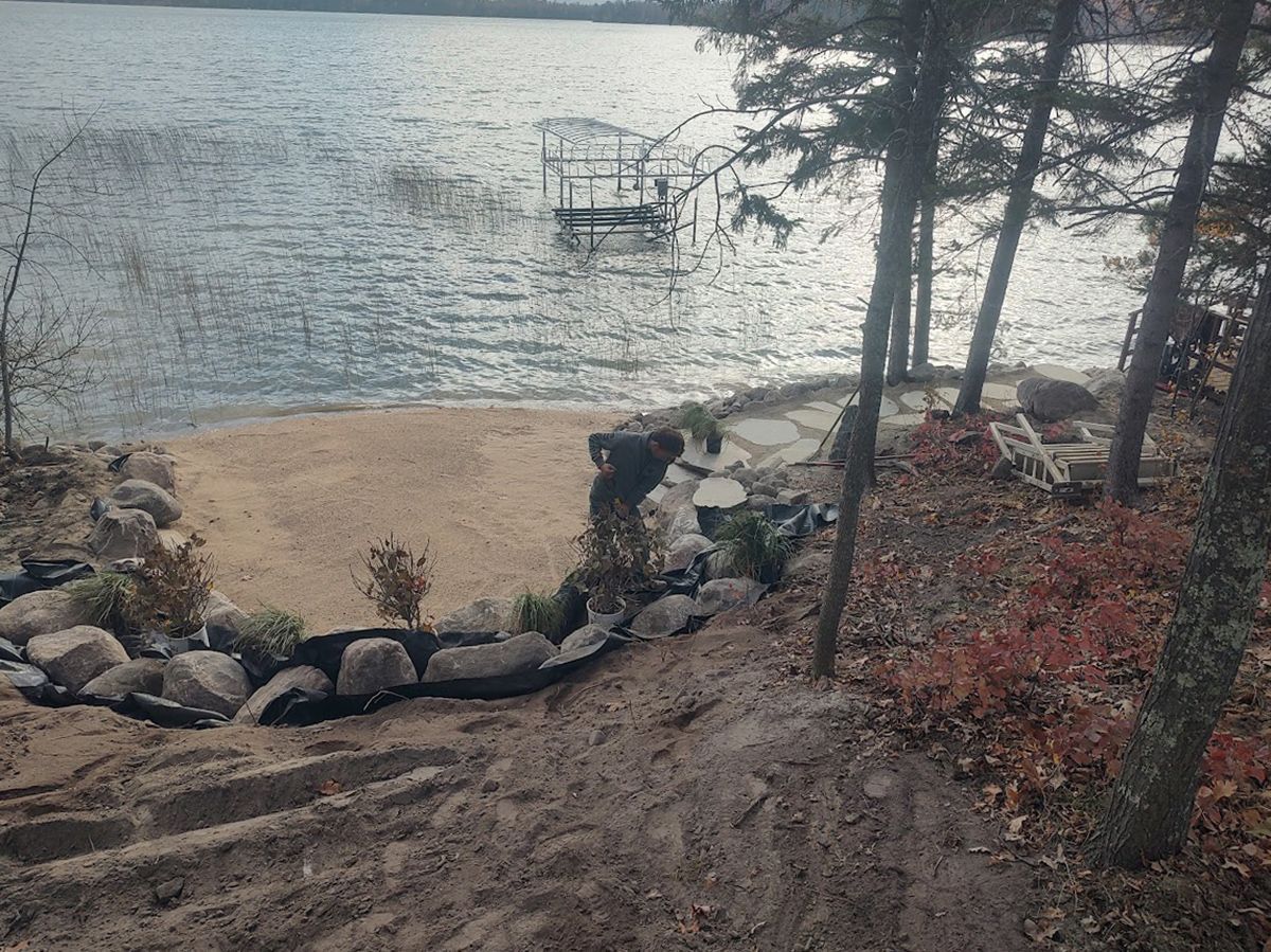 Sandy lakeshore with rocks, trees, and a small dock over calm water