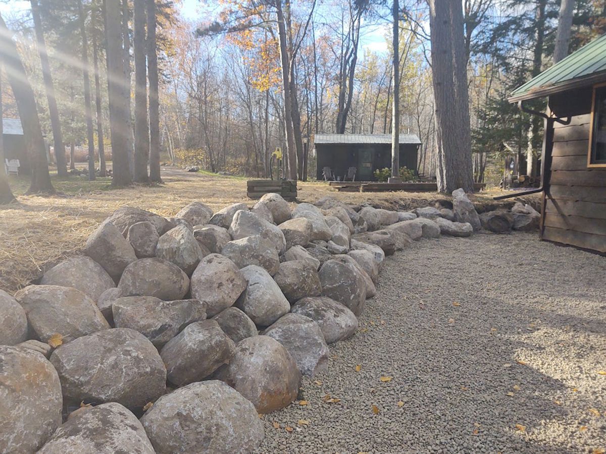 Gravel yard with a large pile of rocks, trees, and a shed in the background beside a building