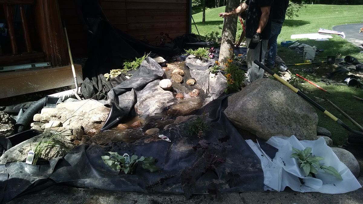 A backyard garden bed covered in black tarp, with rocks, plants, and tools scattered around.