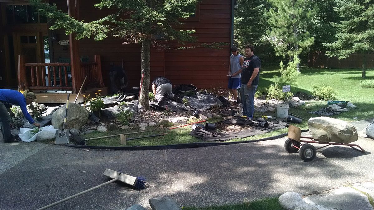 People landscaping outside a wooden house, with wheelbarrows, plants, and yard tools scattered around.