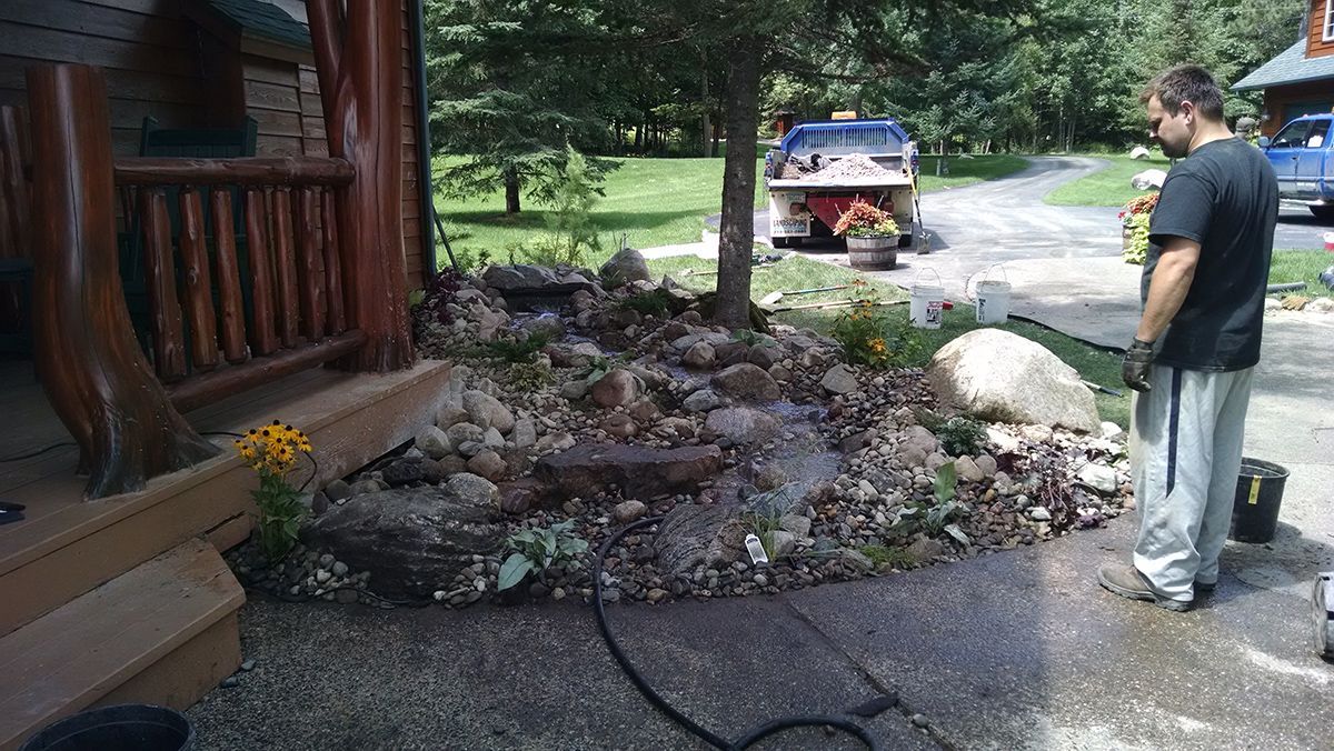 Person standing beside a rocky garden bed next to a cabin, with a truck on a wooded driveway nearby