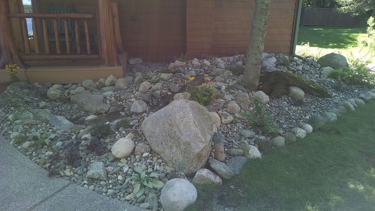 Rock garden with large stones and a palm tree beside a wooden deck, bordered by green grass.
