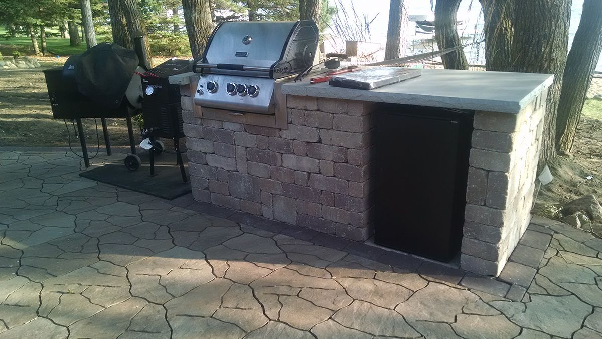 Outdoor brick barbecue grill beside a black smoker on a stone patio in a shaded yard