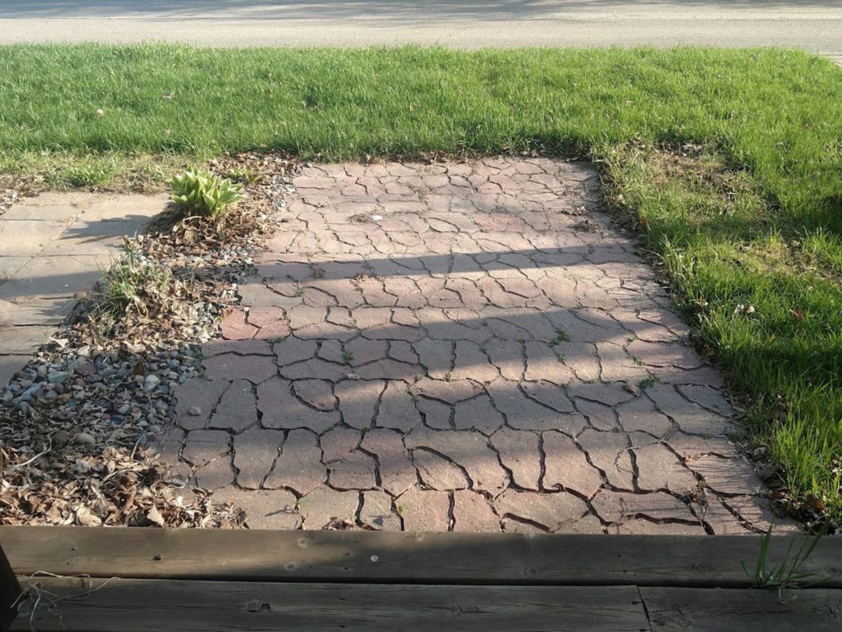 Cracked concrete sidewalk section bordered by grass and fallen leaves in sunlight