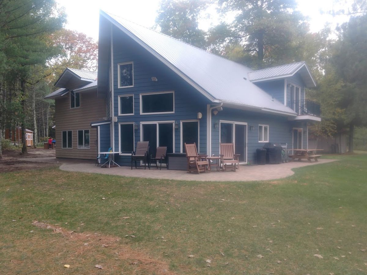 Blue cabin-style house with steep roof and porch chairs in a wooded yard