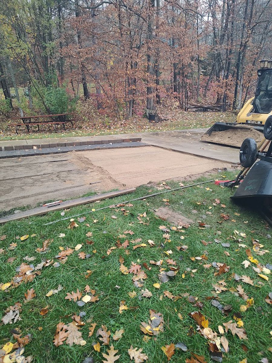 Empty concrete slab beside a grassy yard, with a yellow excavator at the right and autumn woods in back