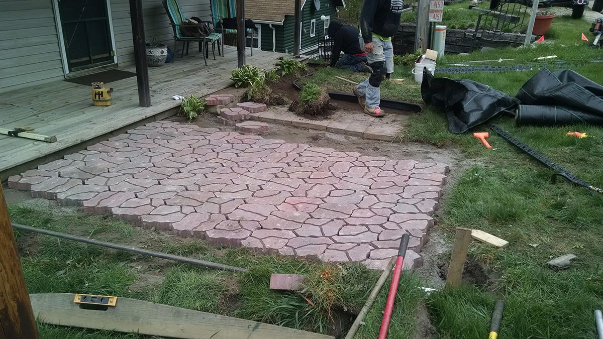 Partially paved backyard with pink brick patio, workers, and stacks of black landscaping material nearby