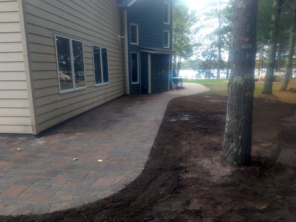 Brick walkway beside a beige house and tree, leading toward a lakeside view.