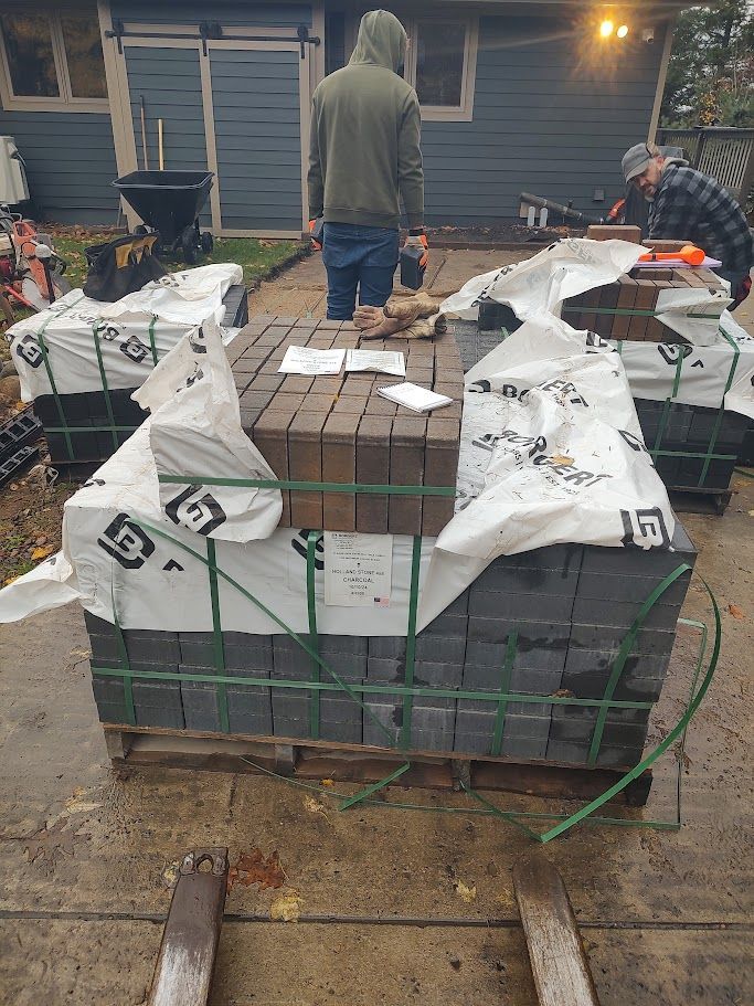 Pallet of stacked pavers on a driveway, wrapped in white plastic, with two workers nearby.