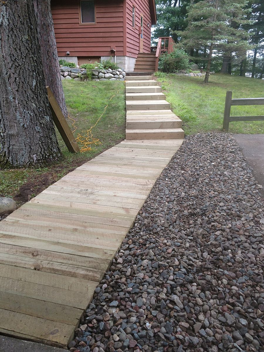 Wooden wheelchair ramp and steps leading to a house with red siding, beside a gravel path and trees.
