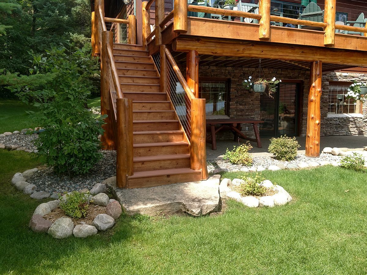 Wooden deck stairs beside a log cabin, with rocks and green lawn below.