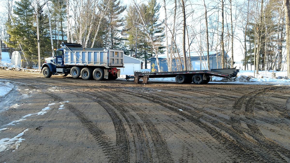 Dump truck hauling a flatbed trailer on a muddy, snow-covered lot with trees in the background