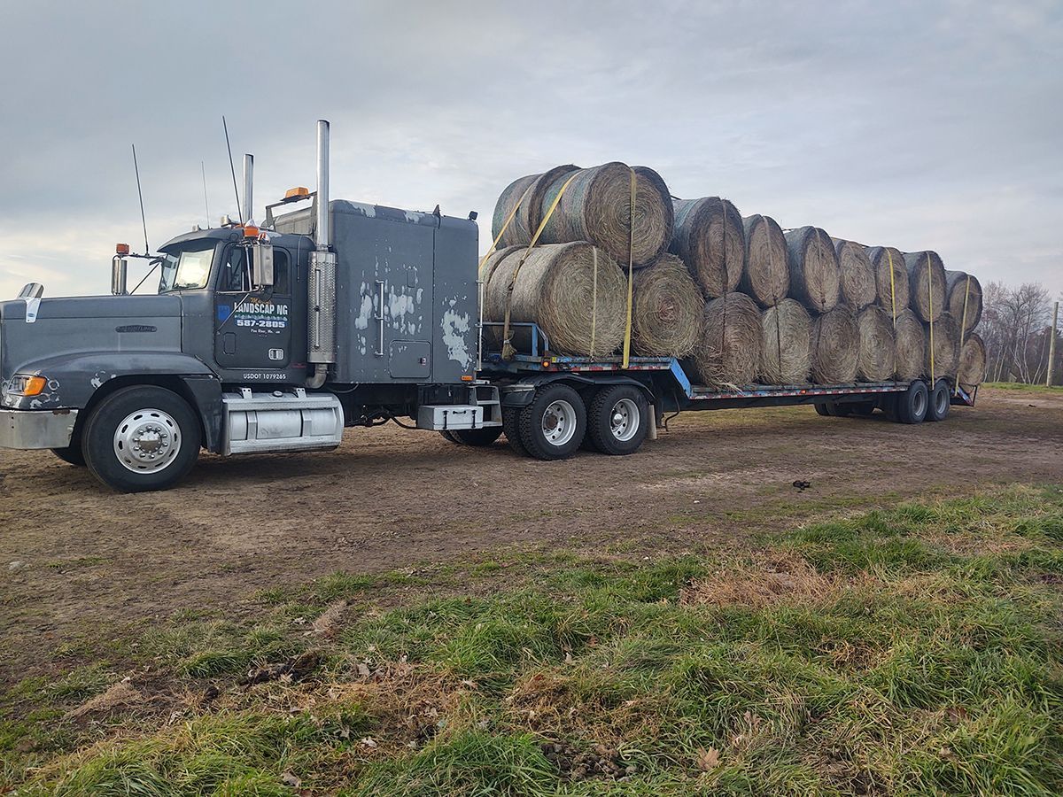 Gray semi-truck hauling a flatbed trailer loaded with large round hay bales on a rural roadside
