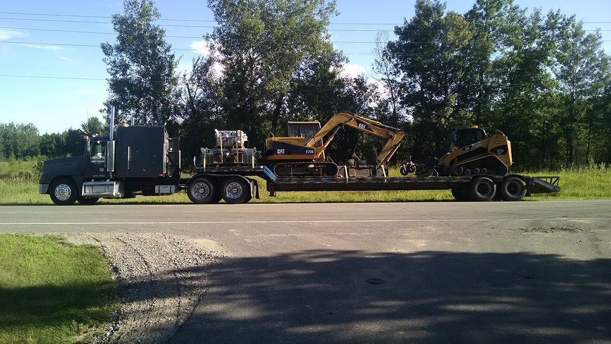 Flatbed truck hauling yellow excavator on a trailer beside a grassy roadside with trees in the background
