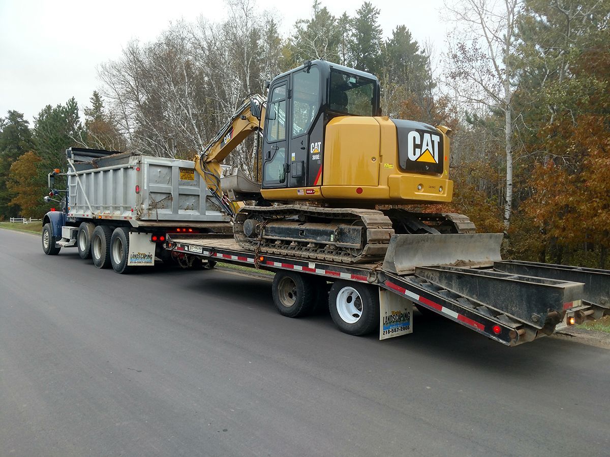Yellow CAT excavator on a flatbed trailer being hauled along a road beside trees.