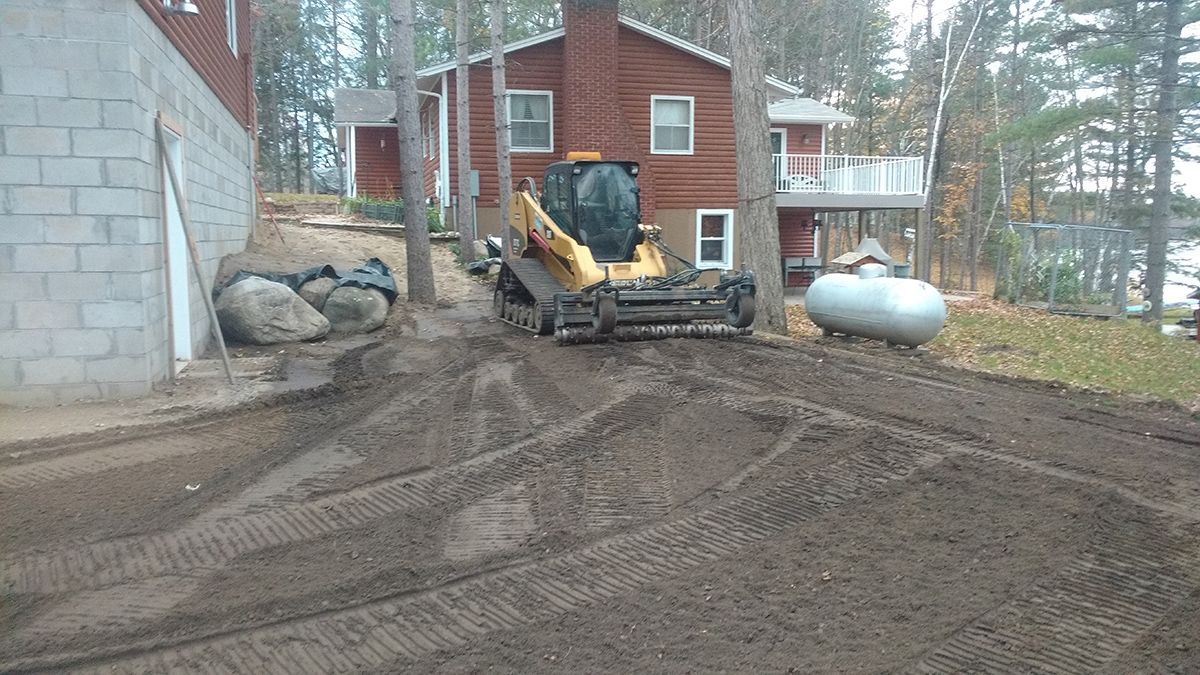 Construction equipment at a muddy house site beside a red brick building and large white tank.