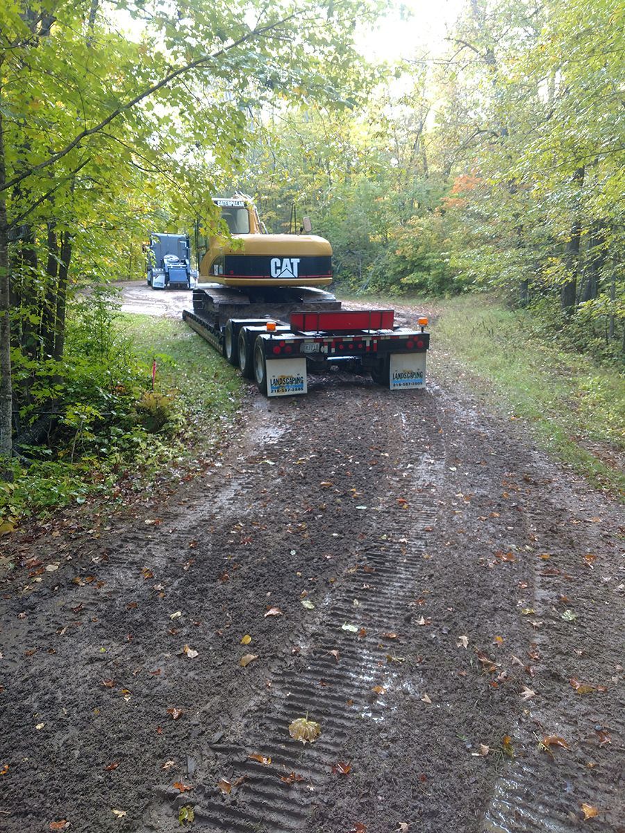 CAT excavator on a flatbed trailer on a wooded dirt road, with a blue truck ahead.