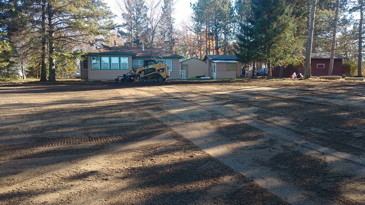 Rural house with large windows among pine trees, viewed across a dirt clearing in late afternoon light