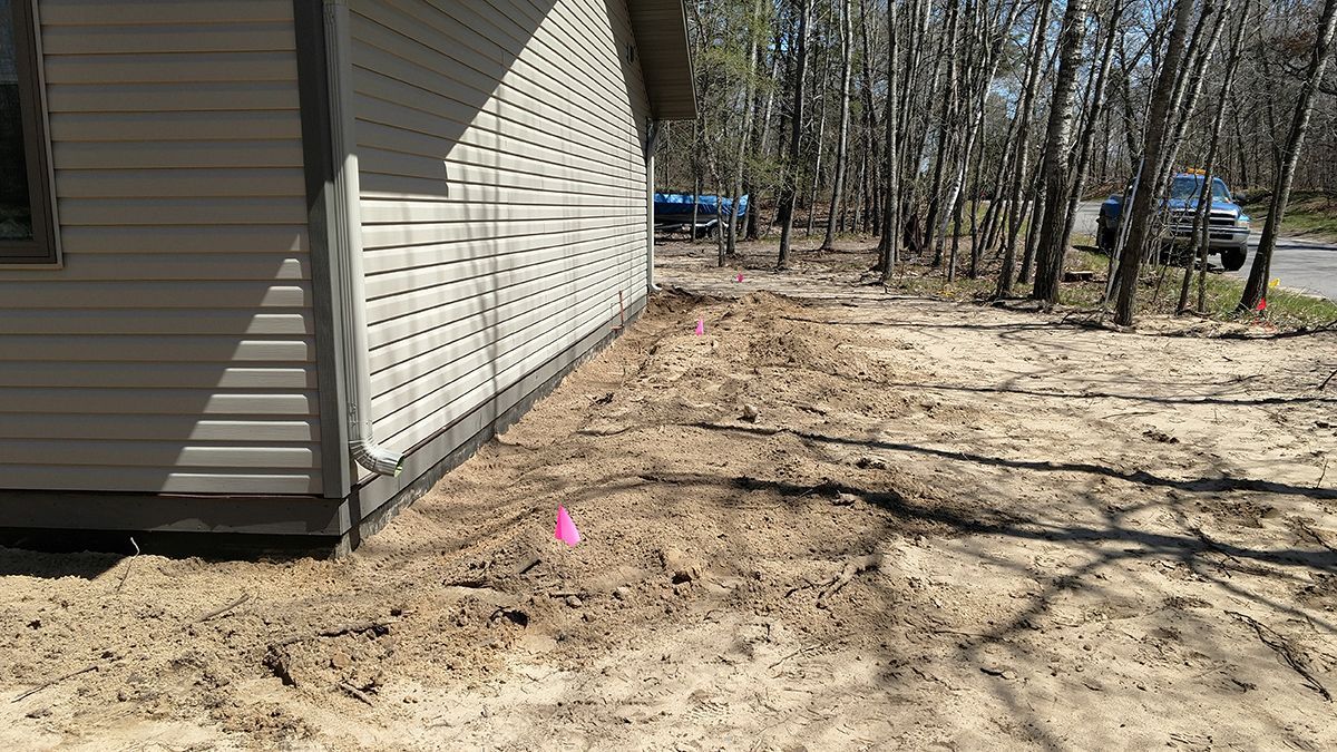 House side yard with bare sandy ground, leafless trees, and a small pink marker near the wall
