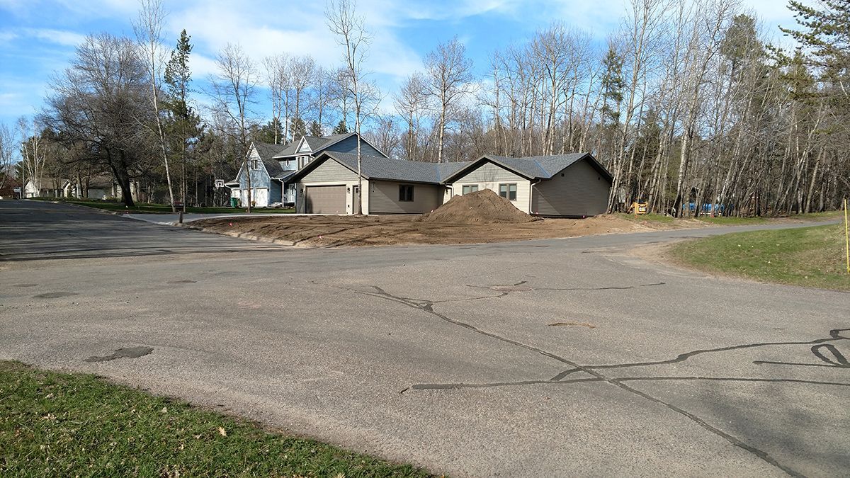 Wide empty parking lot in front of small beige houses and leafless trees under a blue sky.
