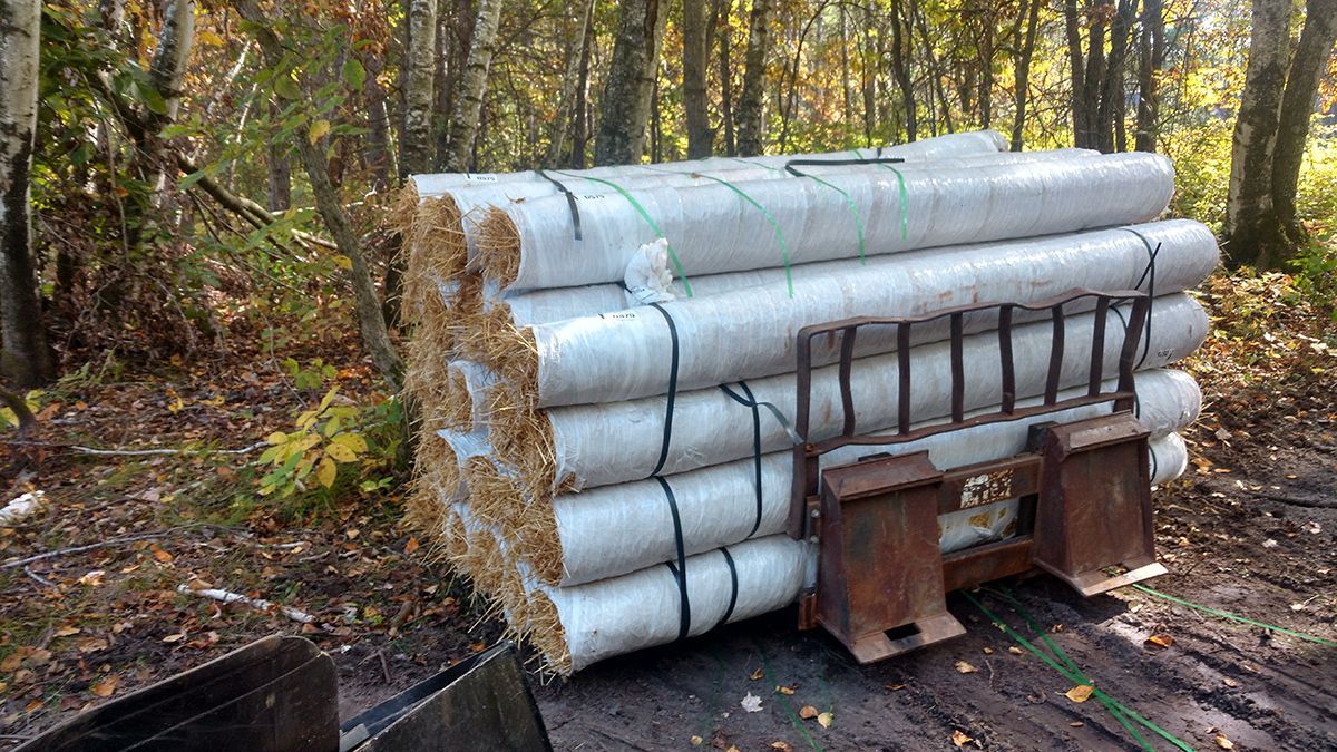 Stacked bundled logs beside a rusty metal splitter in a wooded clearing, with autumn trees in the background