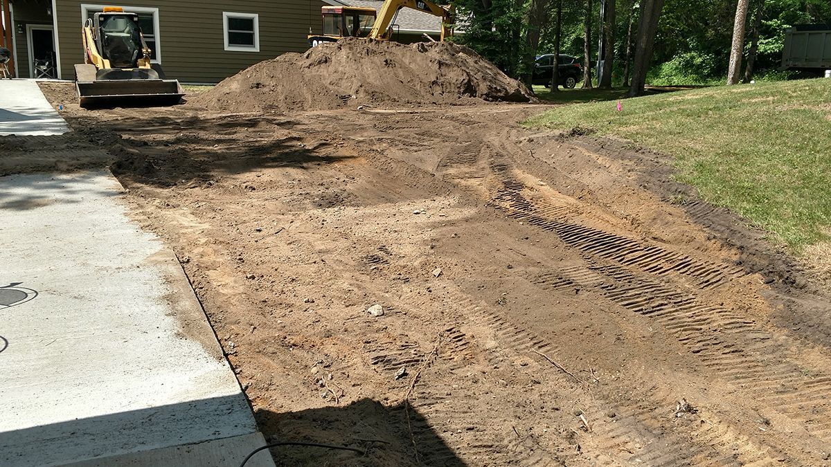 Construction equipment moving dirt beside a driveway, with a large dirt pile and tire tracks in a yard
