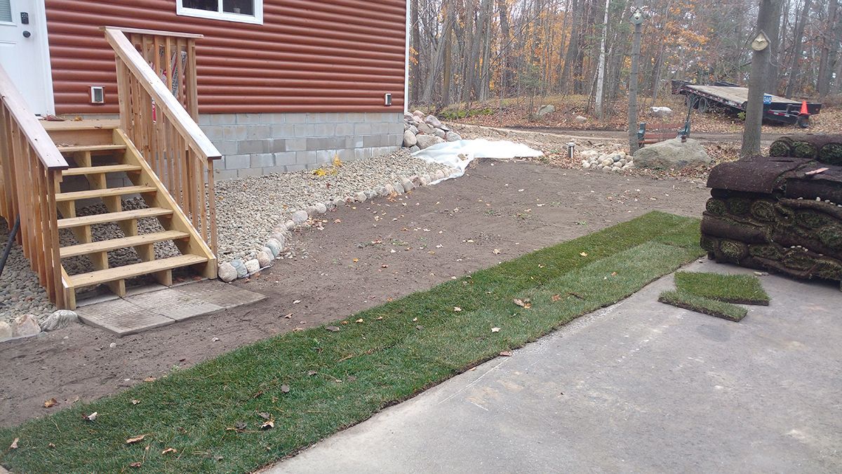 Red house exterior with wooden stairs, gravel yard, and a concrete driveway beside a log pile.