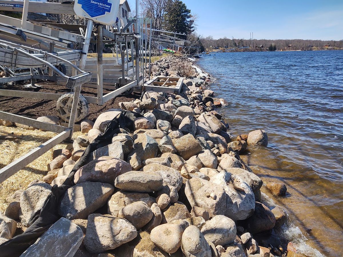 Rock-lined shoreline beside a blue lake with metal railings and trees in the background
