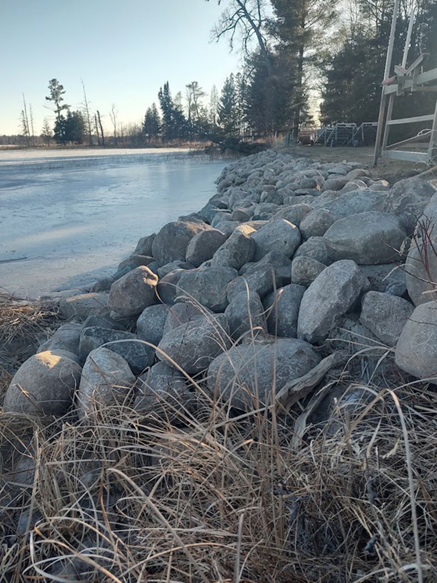 Rocky shoreline beside a frozen lake, with dry grass and trees under a pale winter sky