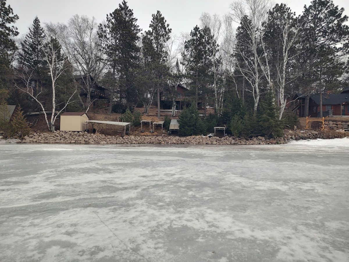 Frozen lake with a snowy shoreline, leafless trees, and cabins in the background