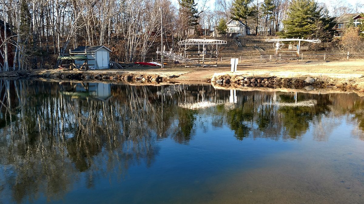 Calm pond reflecting bare trees and a blue sky along a wooded shoreline.