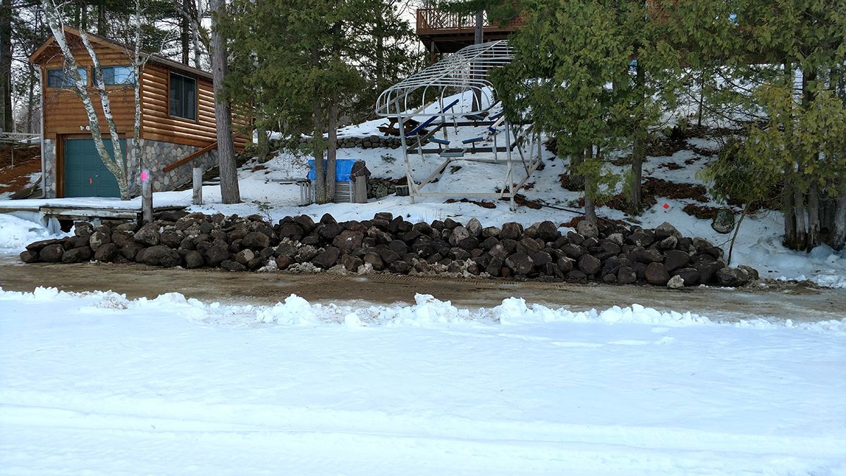 Snowy lakeside cabin with stone wall, pine trees, and frozen water in the foreground