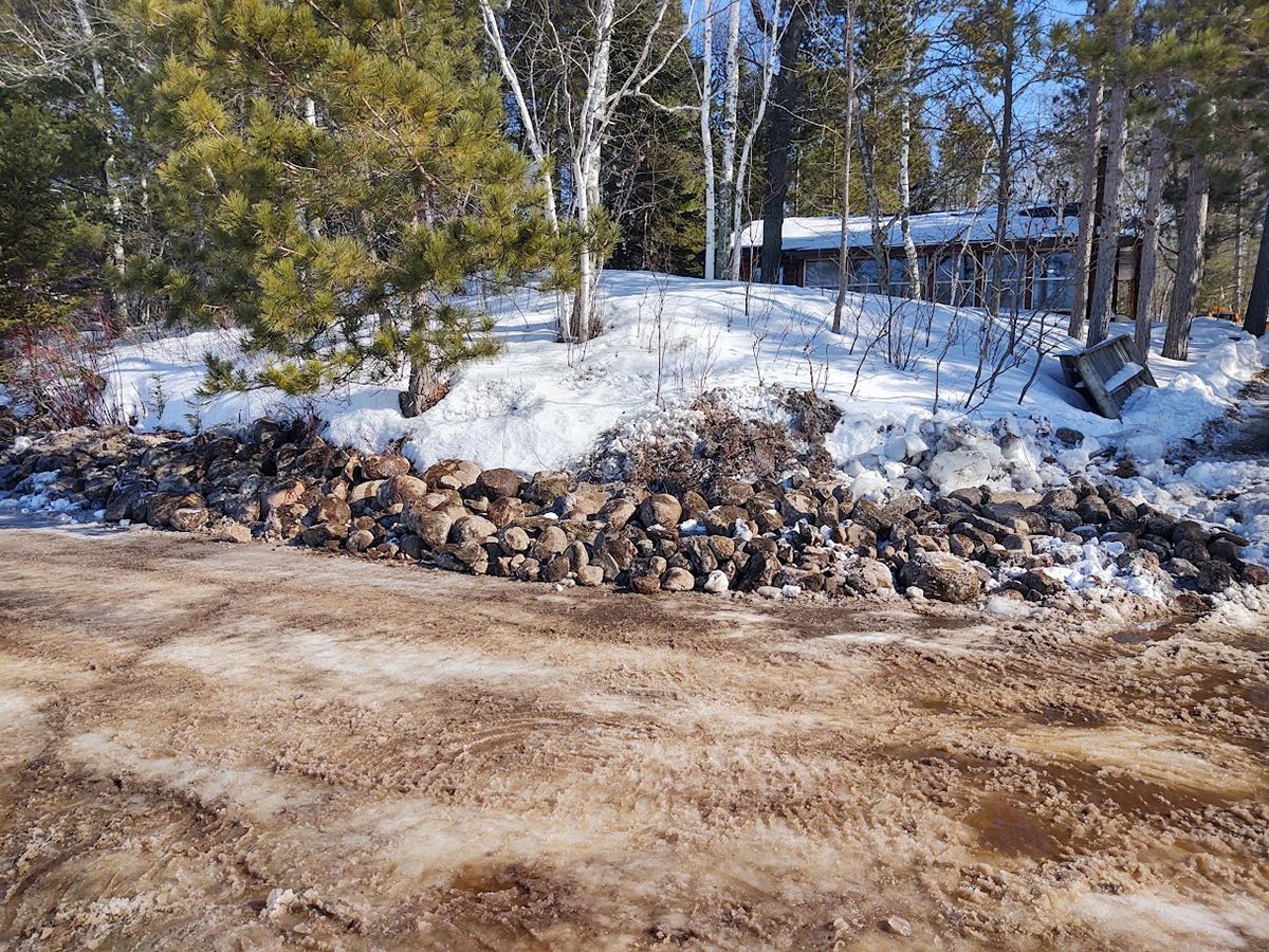Snowy wooded roadside with a stone pile and a cabin in the background