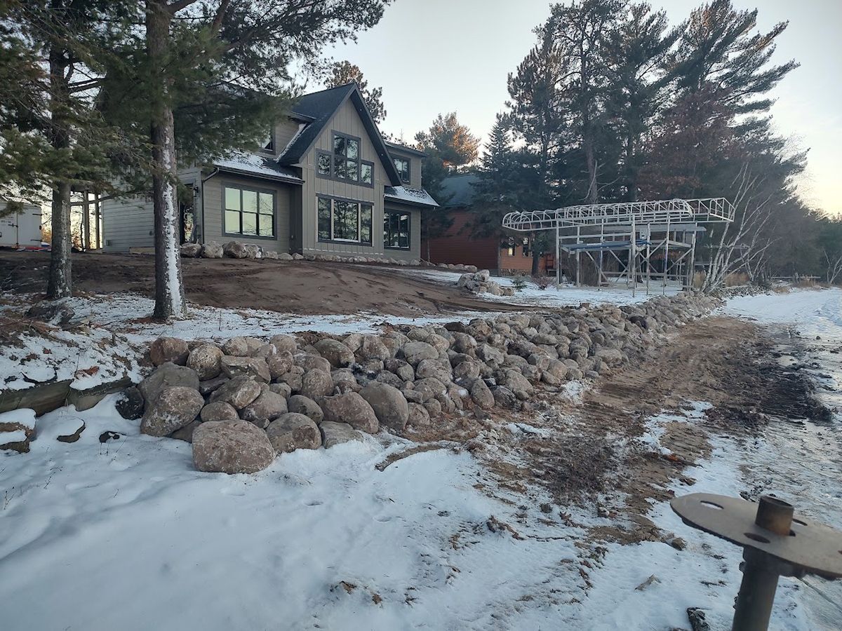 Snowy lakeside house with large windows, rock retaining wall, and pine trees at dusk