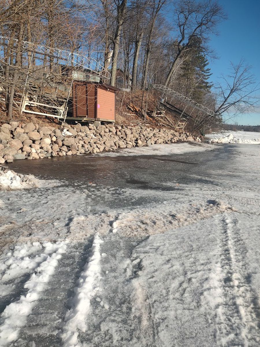 Frozen shoreline with icy water, snow-covered ground, stacked stone embankment, and bare winter trees