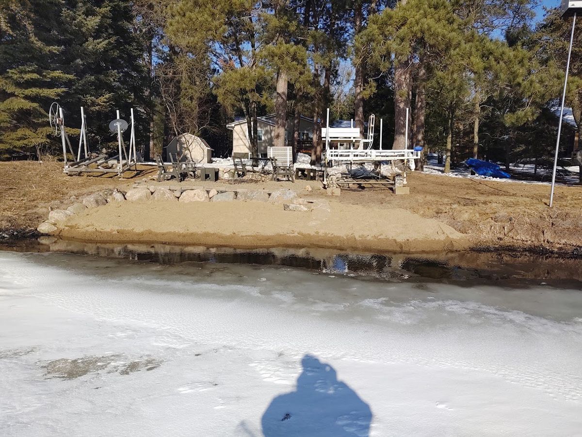 Frozen river with a snowy bank, a small cabin on the far shore, and trees in the background