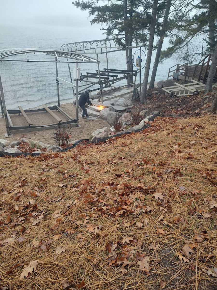 A leaf-covered slope overlooking a lakeside dock with a person near the water under bare trees.
