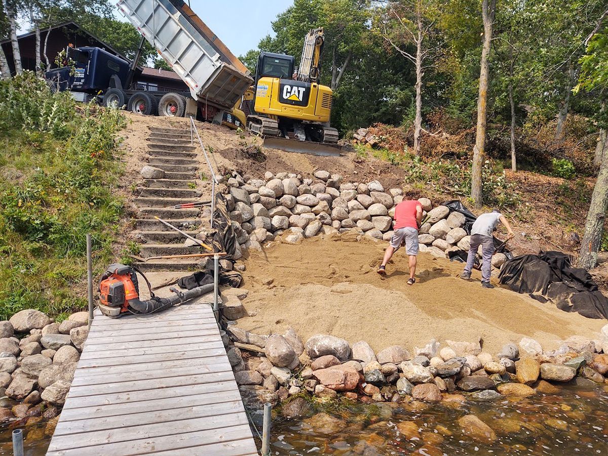 Construction crew building a stone retaining wall beside a stream, with a yellow excavator and wooden bridge nearby