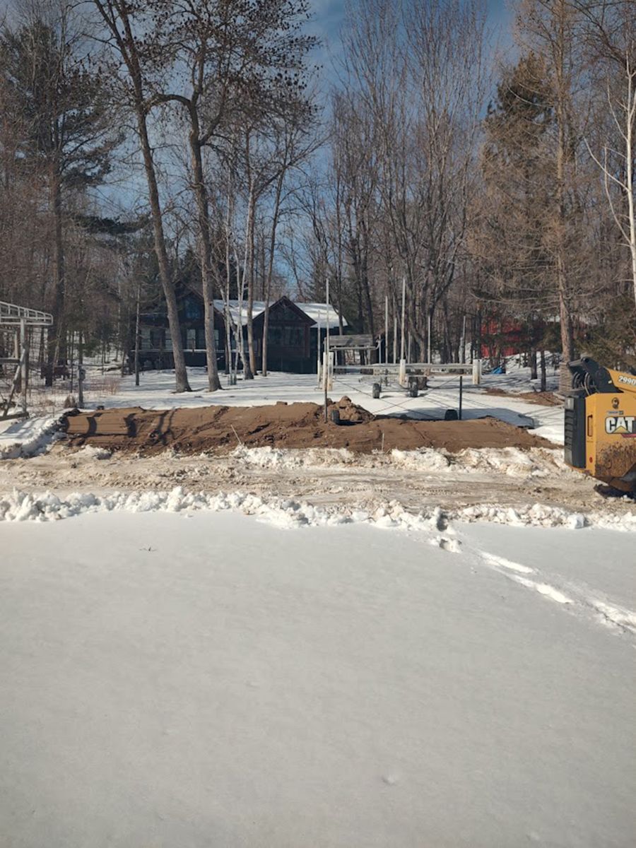 Snowy lakeshore with bare trees, a small cabin, and a yellow excavator near the bank