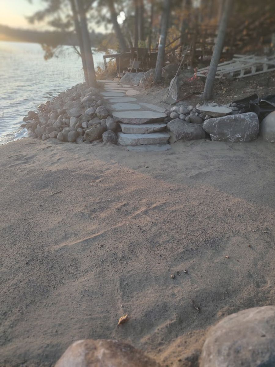 Sandy lakeshore with stone steps, a small rock wall, and trees by the water at sunset