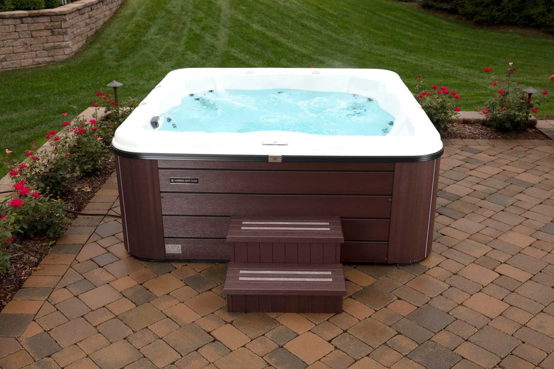 Hot tub with bubbling water on a brick patio, brown exterior, small steps, green lawn in background.