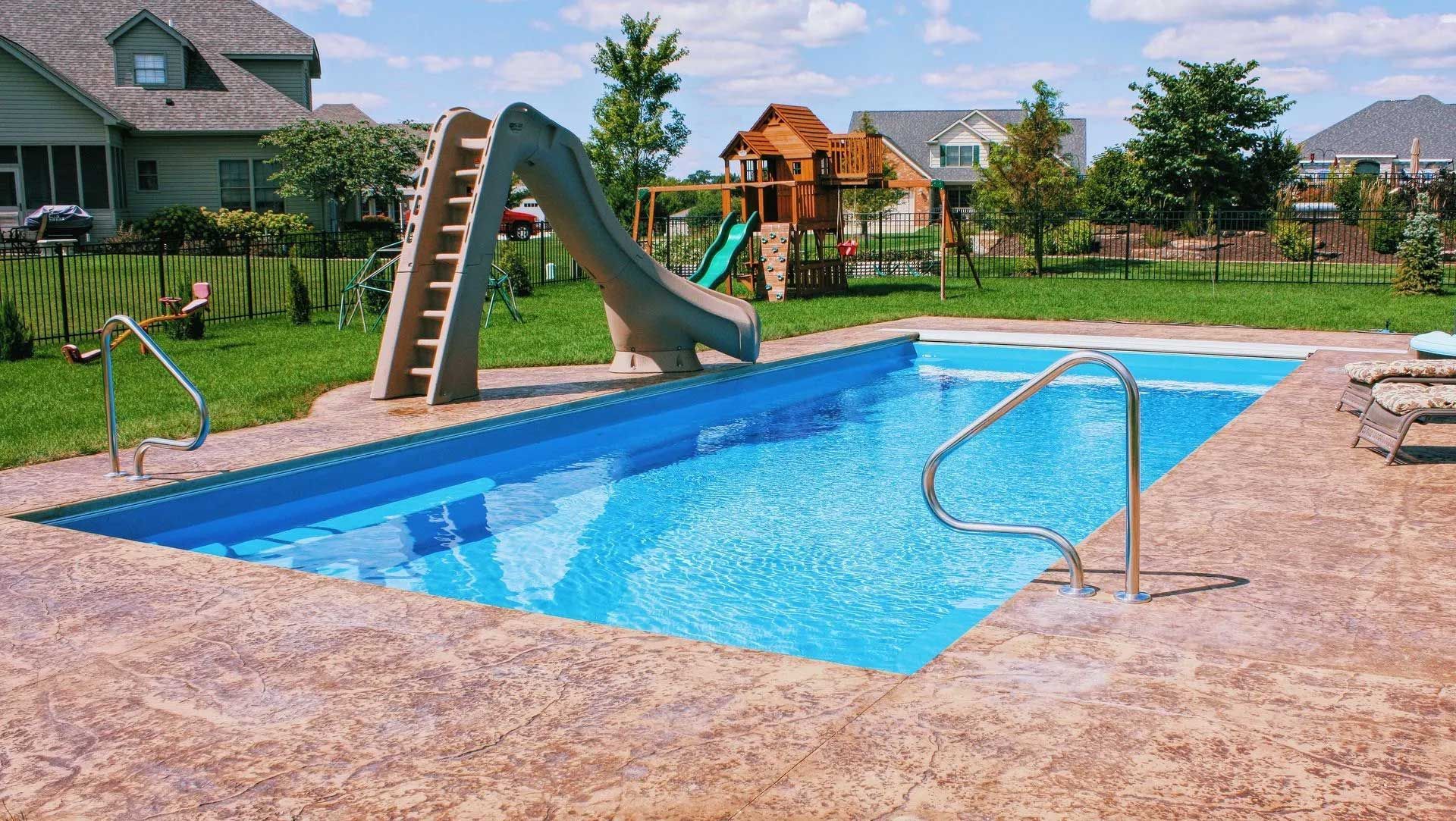 Rectangular in-ground swimming pool with a slide, surrounded by concrete. Backyard with a play structure and houses.