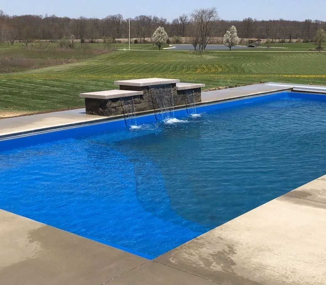 Blue rectangular swimming pool with a stone waterfall feature, set against a green lawn and trees.