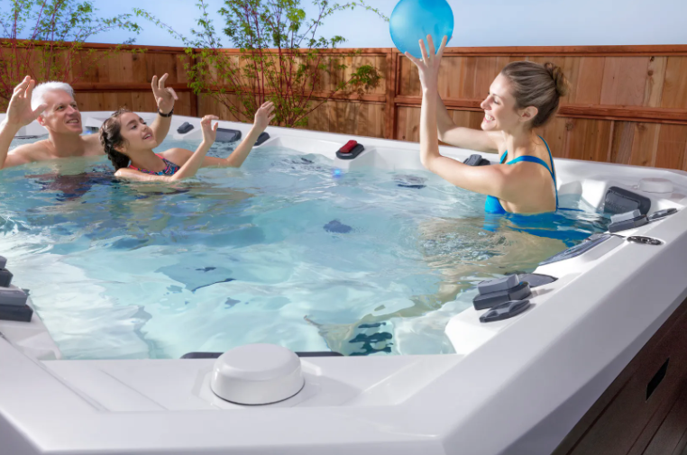 Family playing with a blue ball in a hot tub, outside near a wooden fence.