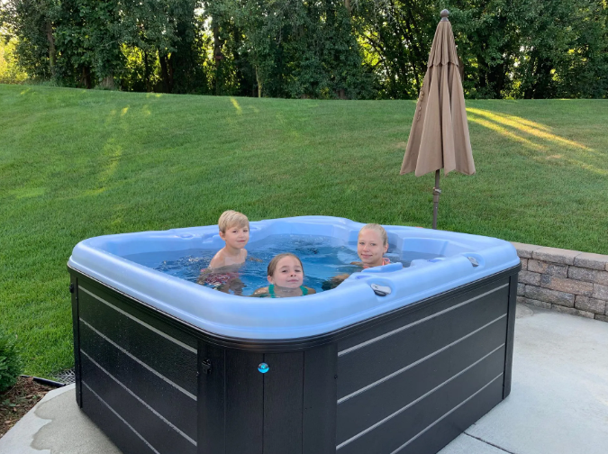 Three children in a square hot tub on a patio. An umbrella shades the tub, green lawn behind.