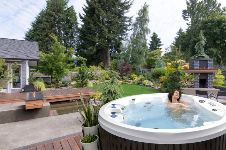 Woman relaxing in a hot tub outdoors. Lush green backyard with trees and a small fireplace.