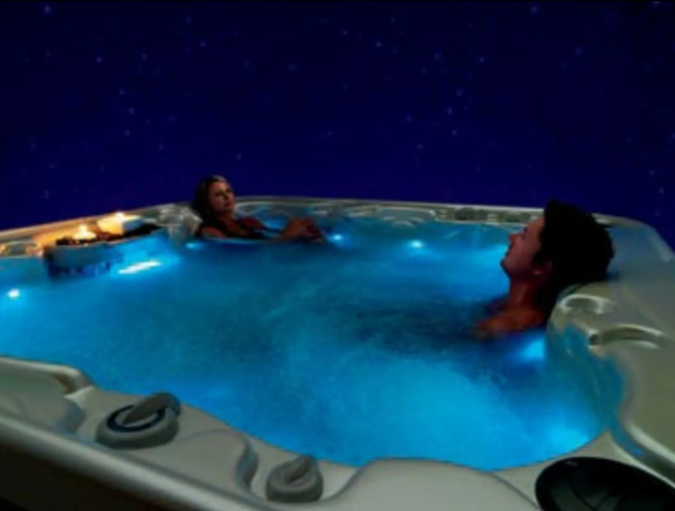 Two people relaxing in a hot tub with blue underwater lights, set against a dark, starry sky.