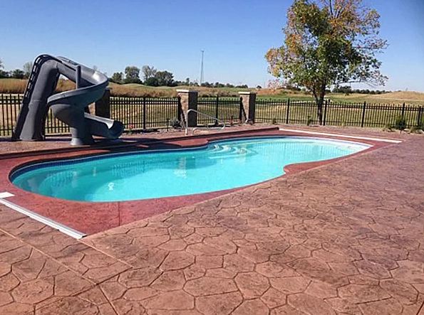 Pool with a slide, surrounded by reddish stamped concrete and a black fence, blue sky.