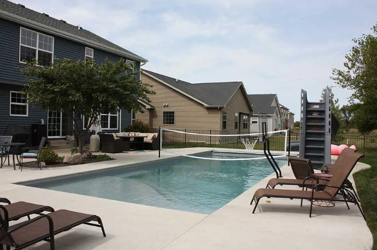 Backyard with pool, loungers, volleyball net, and houses in background on a cloudy day.